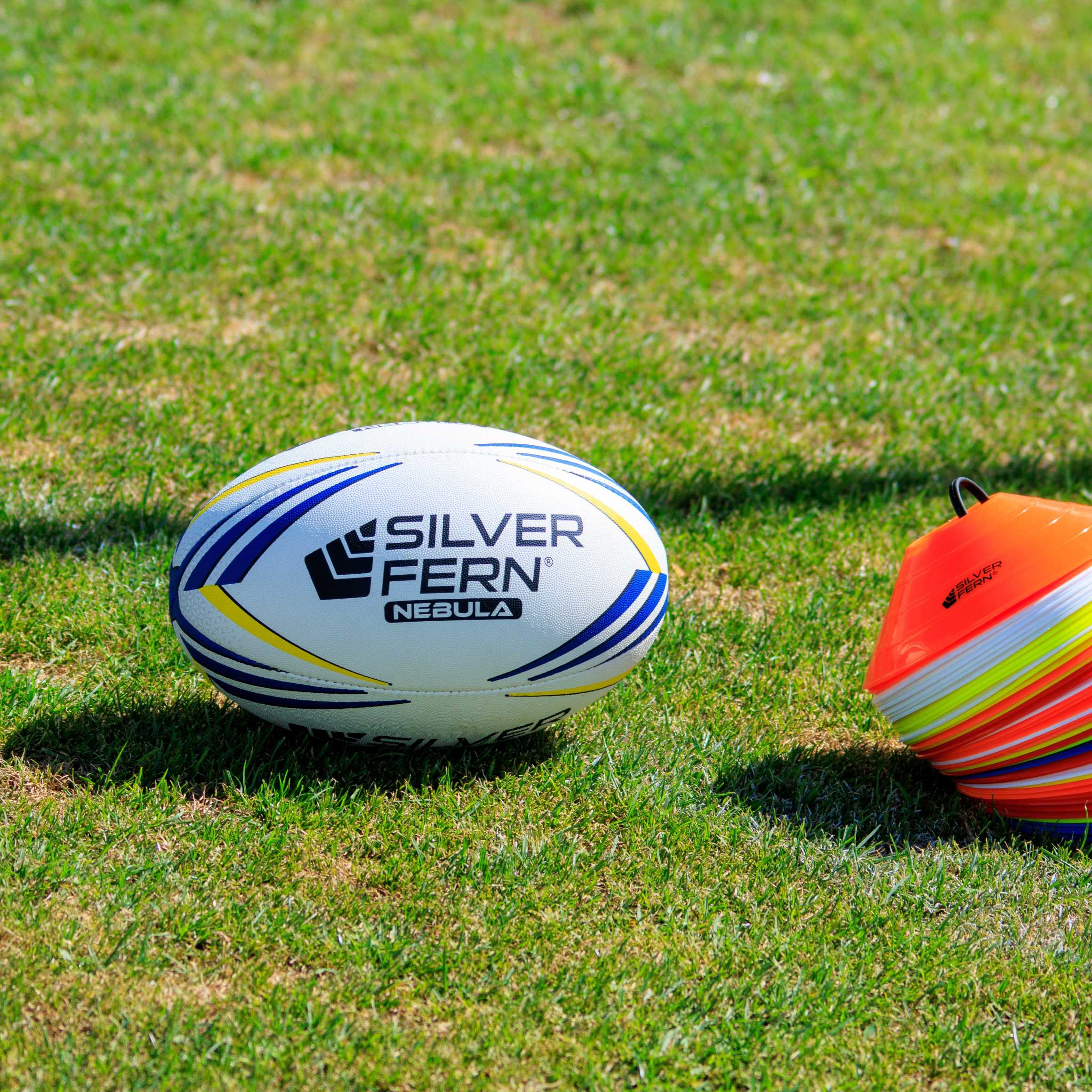 Silver Fern Nebula Premier Match Ball hero shot, sitting on a rugby field next to a stack of marker cones
